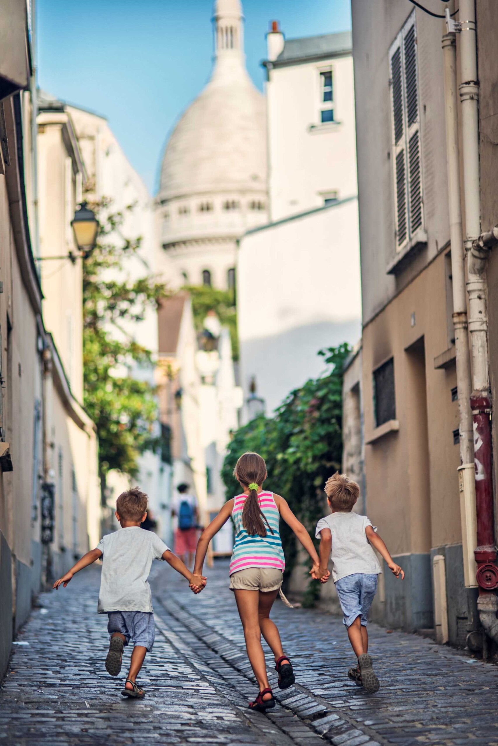 Photo de 3 enfants courant dans les rues de Paris avec le dôme de la Basilique du Sacré-Cœur en fond